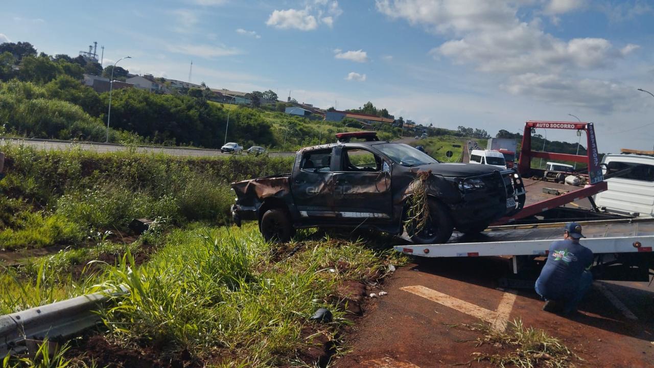 Viatura da Guarda Municipal de Foz do Iguaçu danificada após capotamento na Perimetral Leste, sendo removida por guincho às margens da rodovia, com vegetação ao redor e trânsito ao fundo.
